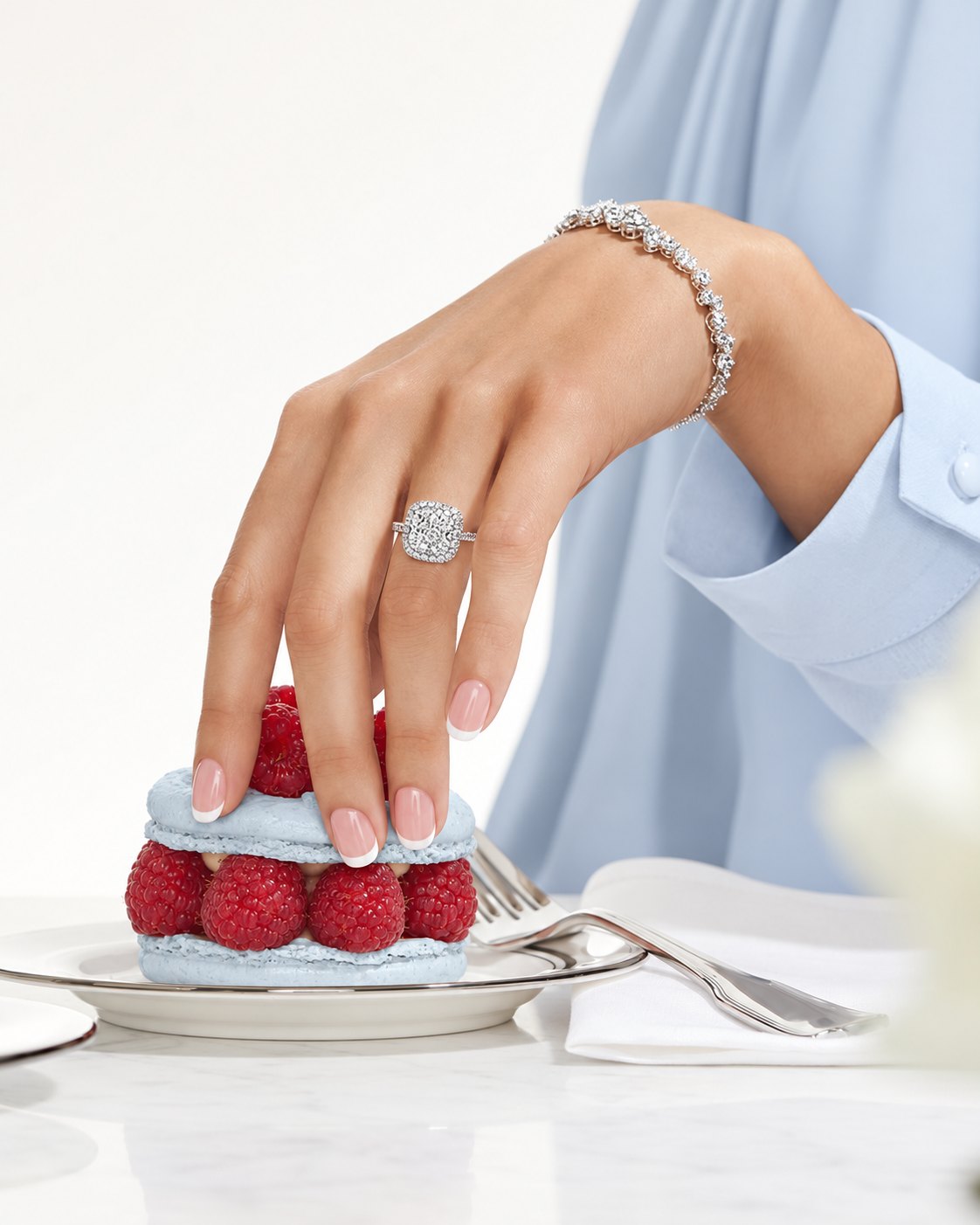 A hand with a diamond ring and bracelet reaching for a powder-blue raspberry macaron